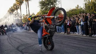 Momento en el que un motorista hace el caballito con una motocicleta de cross en color naranja.