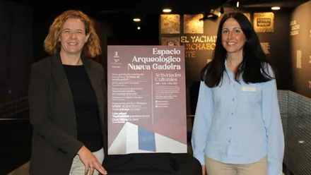 Cartel con las actividades en Nueva Gadeira, la delegada y la responsable posan junto al mismo.