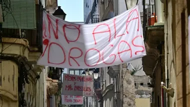 Una gran pancarta blanca con letras rojas y el mensaje "No a las drogas" cuelga entre los edificios de una estrecha calle del barrio de Santa María, en Cádiz. Al fondo se observan otras pancartas reivindicativas, entre ellas una con el lema "Menos papelinas y más papelillos".