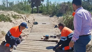 Pasarela de madera de suelo siendo reparada por dos operarios que visten de naranja y azul.