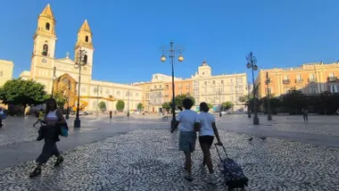 Varias personas caminas por la plaza de San Antonio de Cádiz, con maquinaria de reformas de la iglesia al fondo y un cielo azul
