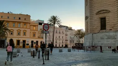 Vista de la plaza de la Catedral de Cádiz al amanecer. Varias personas caminan por el espacio peatonal bajo un cielo despejado, con palmeras y edificios históricos al fondo. La imagen refleja el ambiente suave y templado de una mañana marcada por la primera noche tropical del año en la ciudad.
