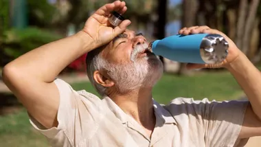Hombre mayor con barba canosa bebe agua mientras le da el sol en un parque.