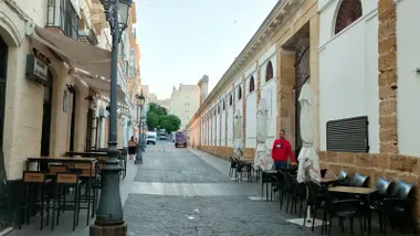 Un hombre coloca sombrillas y mobiliario en la terraza exterior de un bar. El cielo aparece ligeramente velado por la calima y las sombrillas permanecen cerradas debido al viento de Levante. La imagen refleja el ambiente cálido y ventoso que acompaña a la provincia de Cádiz durante este fin de semana.