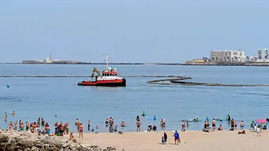 Una draga trabaja frente a la playa de Santa María del Mar mientras decenas de bañistas observan la instalación de las tuberías desde la orilla.