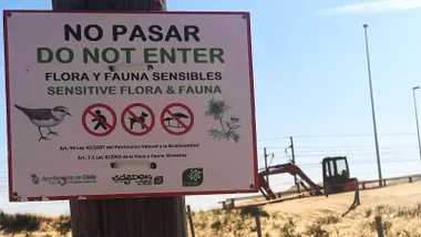 Primer plano de un cartel instalado en las dunas de Cortadura que prohíbe el acceso para proteger la flora y fauna sensibles. Al fondo, sobre la arena, se observa maquinaria utilizada en las obras del trazado ciclista EuroVelo, junto a postes y cuerdas de delimitación. La imagen muestra la coexistencia entre un espacio natural protegido y una infraestructura en construcción o mantenimiento.