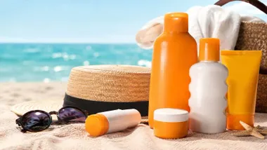 Distintos envases de cremas solares en color blanco y naranja junto a un sombrero de paja en la playa.