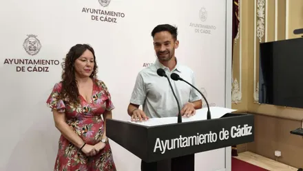 Lorena con un vestido estampado y David de la Cruz con una camisa de color claro, tras ellos el panel del Ayuntamiento de Cádiz en blanco.
