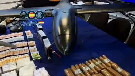 Mesa con fondo azul en el que hay un dron de color negro, fajos de billetes y droga.