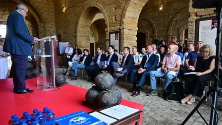 Representantes de la UNESCO, administraciones públicas y organismos de emergencia asisten al acto de entrega de la certificación Tsunami Ready a la ciudad de Cádiz en el interior de la Puerta de Tierra. En primer plano aparece el diploma del reconocimiento y al fondo el público escucha una intervención desde el atril.