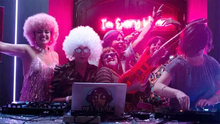 Grupo de mujeres y asistentes con pelucas afro de colores bailando y posando junto a una cabina de DJ iluminada con luces rosas y moradas. Al fondo aparece un letrero luminoso con el nombre I'm Every Woman, en una escena que refleja el ambiente festivo y la estética funk característica del evento.