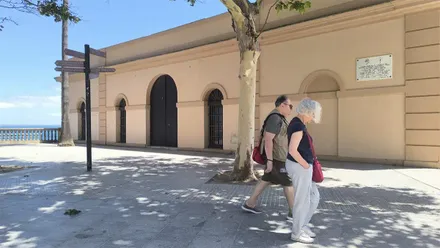 Vista exterior de la Sala La Canela, situada en el Baluarte de la Candelaria. En primer plano, una pareja camina por delante de la fachada de piedra del edificio, junto al acceso al recinto histórico.