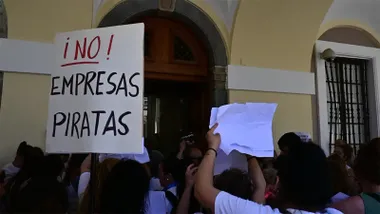 Grupo de trabajadoras del servicio de limpieza de dependencias municipales concentradas ante la puerta principal del Ayuntamiento de Cádiz. Varias personas sostienen una pancarta blanca con el mensaje "No empresas piratas" mientras participan en una protesta relacionada con la futura adjudicación del contrato del servicio. Al fondo se aprecia la puerta del edificio consistorial.
