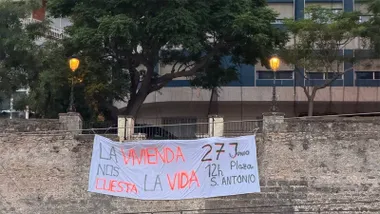 Gran pancarta blanca colgada sobre una muralla de Cádiz con el mensaje “La vivienda nos cuesta la vida” y la convocatoria de una manifestación el 27 de junio en la Plaza San Antonio. Al fondo se observa un edificio de varias plantas con franjas azules y blancas y varios árboles.