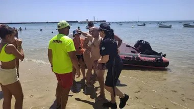 En la orilla de la playa de La Caleta, varias personas rodean a un hombre recién rescatado del mar. Junto a él se encuentran una agente de la Policía Local y un socorrista con camiseta amarilla fluorescente. Detrás aparece una embarcación neumática utilizada en el operativo de rescate. Al fondo se observan bañistas, pequeñas embarcaciones fondeadas y el perfil del castillo de San Sebastián bajo un cielo despejado.