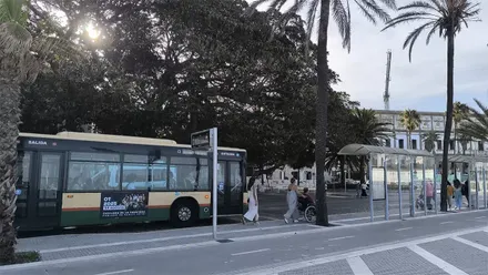 Autobús urbano de Cádiz entrando en la parada de La Caleta mientras varios pasajeros esperan en la acera. Al fondo se distinguen el ficus del Mora y parte del edificio de Valcárcel en una jornada soleada.