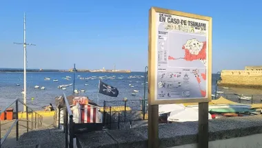 Una bandera de un kiosco de helados se mueve por el Levante en la playa de la Caleta, con un cartel del protocolo por tusamis en primer plano y el castillo de San Sebastián al fondo