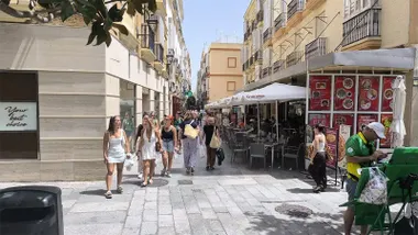 Calle peatonal del centro de Cádiz con varias terrazas de bares, comercios abiertos y numerosos viandantes caminando en una jornada de verano.