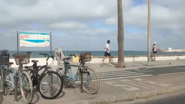 Tres bicicletas aparcadas en el paseo marítimo de Cádiz por el que corre una persona. En el horizonte se ven algunas nubes
