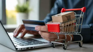 Persona comprando online, con un ordenador, tarjeta en mano y carrito a su lado con cajas.