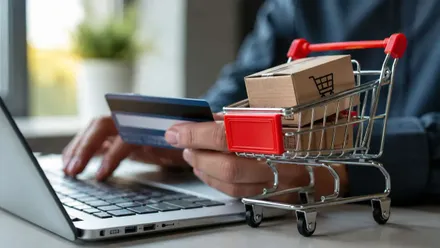 Persona comprando online, con un ordenador, tarjeta en mano y carrito a su lado con cajas.