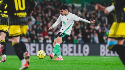 Damián Rodríguez golpea el balón con la pierna derecha durante un partido del Racing de Santander. El centrocampista viste la camiseta blanca con detalles verdes mientras varios jugadores rivales, de espaldas y con equipación negra y amarilla, rodean la acción en un estadio lleno de público.