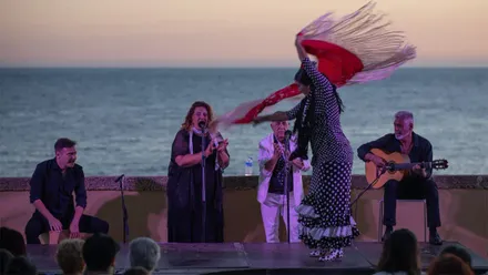 Una bailaora realiza un movimiento con un mantón rojo sobre un escenario al aire libre mientras una cantaora, un guitarrista y un percusionista actúan frente al mar al atardecer, durante una actuación de El Faro Flamenco en el Baluarte de los Mártires de Cádiz.