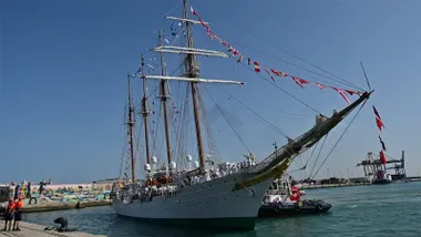 El buque escuela Juan Sebastián de Elcano entra en el puerto de Cádiz engalanado con banderas. En cubierta, decenas de guardiamarinas uniformados permanecen formados mientras un remolcador acompaña la maniobra de atraque bajo un cielo completamente despejado.