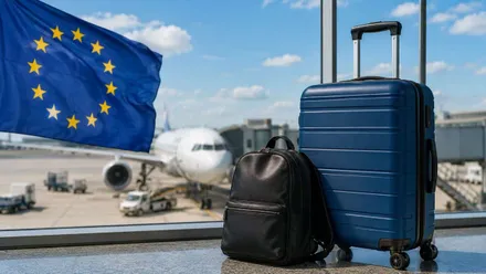 Maleta de mano y bolso, junto a un ventanal desde el que se ve un avión y la bandera de la UE (Unión Europea).