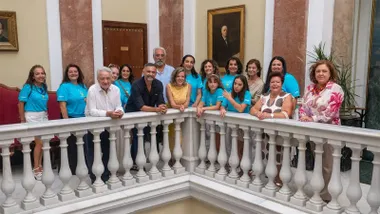 Fotografía de grupo tomada en el patio del Ayuntamiento. Quince personas, entre representantes municipales, organizadores y participantes del Festival Internacional de Folklore 'Ciudad de Cádiz', posan apoyadas sobre una barandilla de mármol. Varias de ellas visten camisetas turquesa con la imagen del festival, mientras otras llevan ropa de calle. Al fondo se observan columnas de mármol, retratos enmarcados y elementos arquitectónicos clásicos del edificio donde tuvo lugar la presentación oficial.