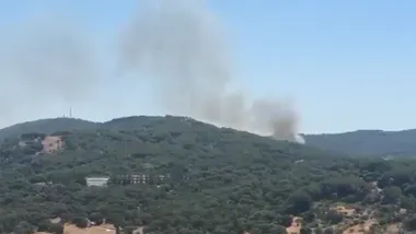 Vista panorámica de una zona de monte en Grazalema con una densa columna de humo gris que se eleva desde detrás de una ladera cubierta de vegetación. En primer plano se distinguen varios edificios entre los árboles y el cielo aparece completamente despejado.