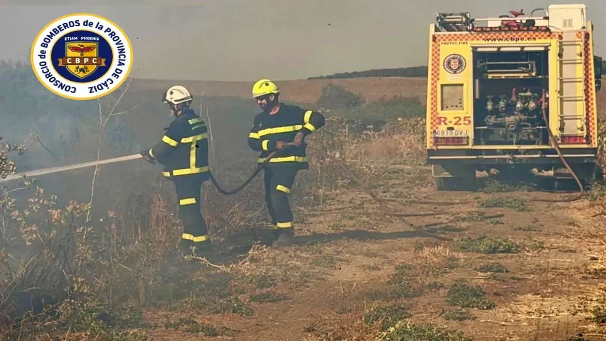 Camión de bomberos y dos bomberos apagando un incendio en zona de pastos.