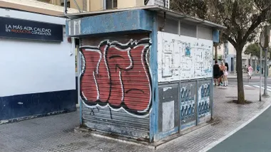 Fotografía de un quiosco metálico cerrado con la persiana bajada y pintada con un gran grafiti en tonos rojos y negros. La estructura, de color azul y con carteles deteriorados en los laterales, ocupa parte de una acera amplia en Cádiz. A la derecha hay un árbol, un paso de peatones y varias personas caminando o conversando al fondo.
