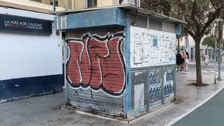 Fotografía de un quiosco metálico cerrado con la persiana bajada y pintada con un gran grafiti en tonos rojos y negros. La estructura, de color azul y con carteles deteriorados en los laterales, ocupa parte de una acera amplia en Cádiz. A la derecha hay un árbol, un paso de peatones y varias personas caminando o conversando al fondo.