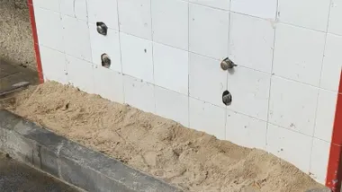 Primer plano de un módulo de lavapiés en una playa de Cádiz. La pared está revestida con azulejos blancos y varios grifos aparecen desmontados o inutilizados. En la parte inferior se acumula una gran cantidad de arena sobre la plataforma de hormigón, sin que se aprecie agua en funcionamiento.