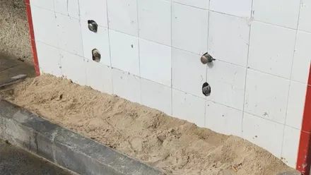Primer plano de un módulo de lavapiés en una playa de Cádiz. La pared está revestida con azulejos blancos y varios grifos aparecen desmontados o inutilizados. En la parte inferior se acumula una gran cantidad de arena sobre la plataforma de hormigón, sin que se aprecie agua en funcionamiento.