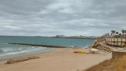 Vista de la playa de Santa María del Mar, en Cádiz, con el cielo cubierto por abundantes nubes bajas, el mar ligeramente agitado y la arena prácticamente vacía. Al fondo se aprecia el perfil de la ciudad, reflejando el ambiente fresco y nuboso tras el cambio de tiempo.
