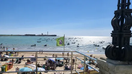 La badera verde del estado del mar en la playa de la Caleta se muevecon el viento de Poniente. AL fondo se ve a contraluz el castilllo de San Sebastián