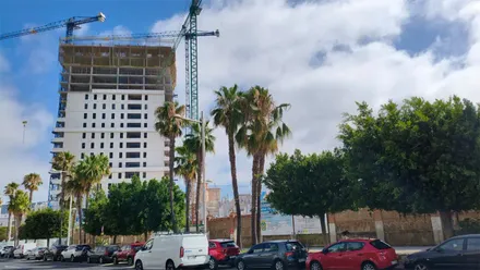 Obras de una torre de viviendas en la avenida de Marconi de Cádiz, con la estructura en construcción, dos grúas, palmeras y coches aparcados en primer plano.