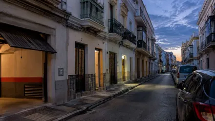 Calle con algunas casas iluminadas y el atardecer cayendo. Hay coches aparcados a la izquierda.