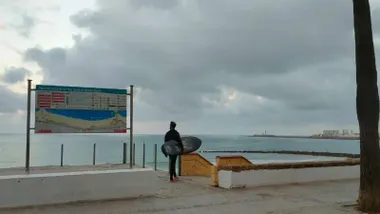 un hombre con una tabla de surf se prepara para bajar a la playa de Santa María del Mar de Cádiz. Junto a él, el cartel informativo de dicho arenal y al fondo, nubes en el horizonte
