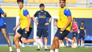 Albert Celades, de pie y con las manos en la cintura, observa un ejercicio durante un entrenamiento del Cádiz CF sobre el césped del Nuevo Mirandilla. Varios jugadores entrenan a su alrededor con petos amarillos.