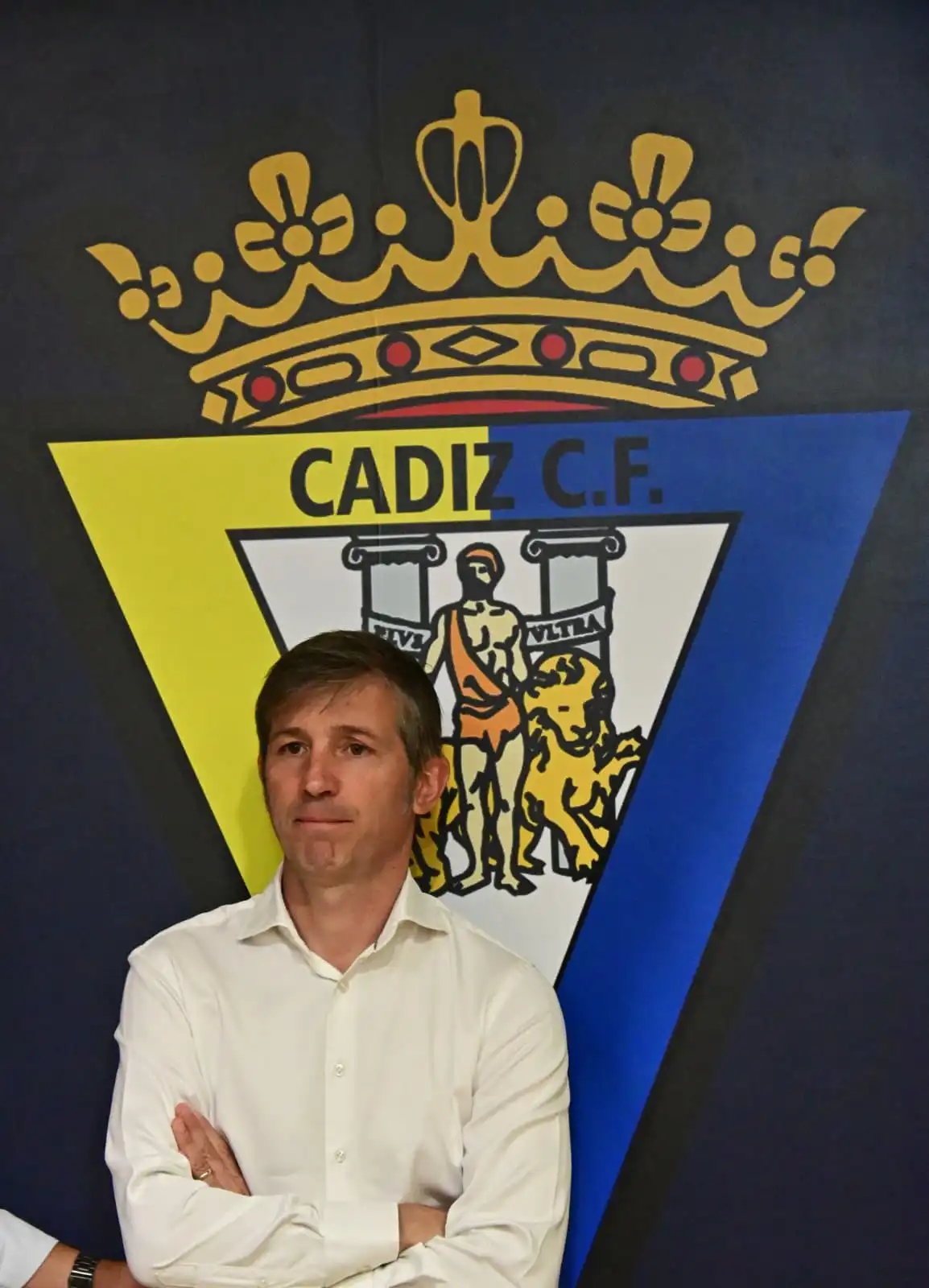 Albert Celades sigue la rueda de prensa de Christian Septien y rafael Contreras mientras espera su turno para ser presentado apoyado en un escudo del Cádiz CF pintado en la pared. Foto: Eulogio García.