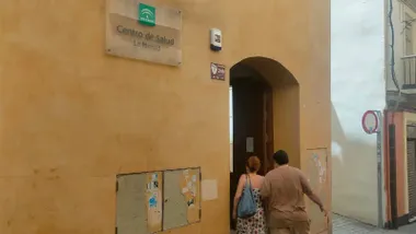 Fachada del Centro de Salud La Merced de Cádiz. Dos personas entran por la puerta principal del edificio, sobre la que se encuentra el cartel identificativo del centro sanitario.