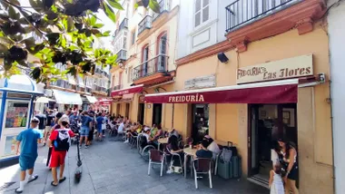 Fachada de la Freiduría Las Flores en una concurrida calle del centro de Cádiz. Varias personas ocupan las mesas de la terraza y pasean frente al establecimiento, identificado por sus toldos granates y el rótulo de Las Flores.