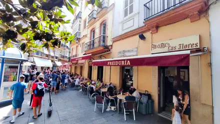 Fachada de la Freiduría Las Flores en una concurrida calle del centro de Cádiz. Varias personas ocupan las mesas de la terraza y pasean frente al establecimiento, identificado por sus toldos granates y el rótulo de Las Flores.