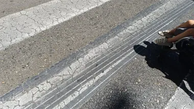 Varias marcas oscuras y alargadas de neumáticos atraviesan la calzada junto a un paso de peatones en la entrada a Cádiz.