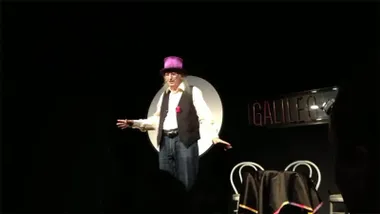 Juan Tamariz actúa sobre un escenario vestido con camisa blanca, chaleco negro y sombrero morado, frente a una mesa de magia y con un fondo oscuro.