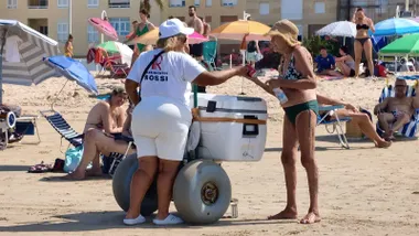 Una vendedora ambulante vestida de blanco atiende a una bañista junto a un carro con una gran nevera, mientras numerosos usuarios descansan bajo sombrillas y en sillas sobre la arena.