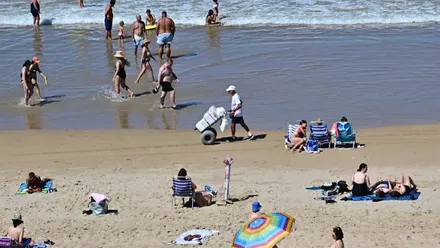 Vista de una playa de Cádiz llena de bañistas. En el centro de la imagen, un vendedor ambulante vestido con camiseta y gorra blancas camina por la orilla mientras arrastra un carro cargado con productos.